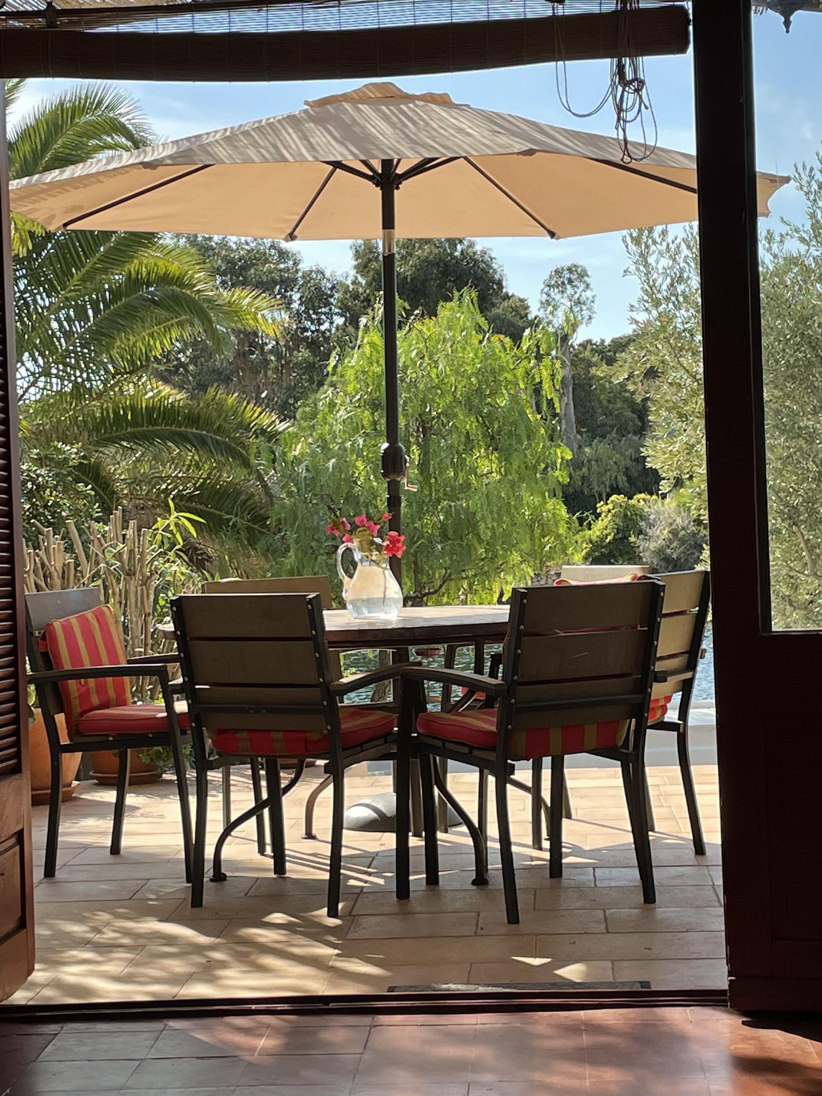 Une terrasse extérieure avec une table et des chaises, un grand parasol et une vue sur un jardin avec des arbres et une étendue d’eau.
