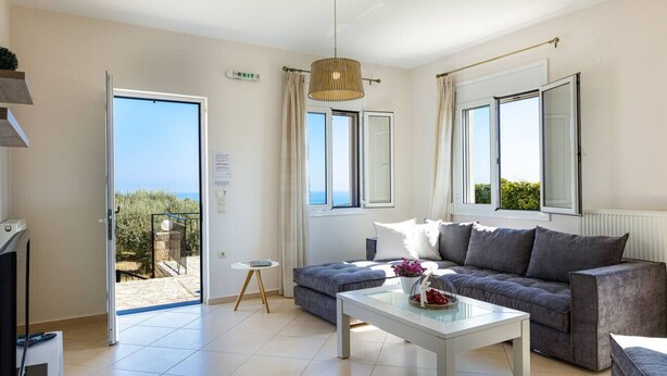 Un salon moderne avec un canapé gris, une table basse blanche et une vue sur la mer grâce à de grandes fenêtres.
