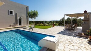Une piscine avec une terrasse en pierre, un espace salon extérieur couvert et une vue sur un paysage verdoyant.