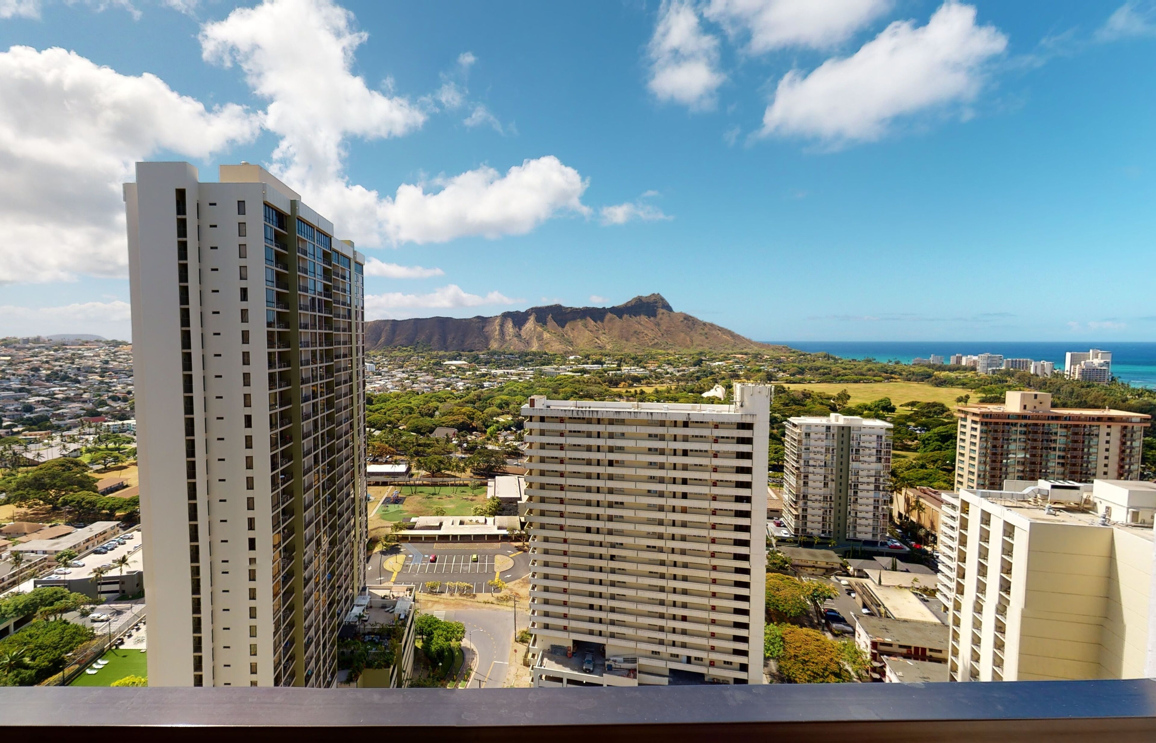 A high-rise building with multiple floors and balconies, surrounded by other buildings and a mountainous landscape in the background.