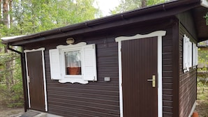 A small, single-story wooden cabin with a brown exterior and white trim. It features a window with shutters, a door, and a small porch area. - Eastern Nostalgia in the Cabin Village (exclusive use) - Yacht tour starting at 7 days, booking included (Groß Köris)