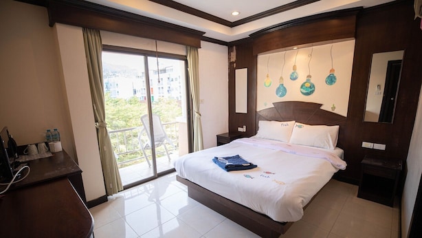 A hotel room with a large bed, a desk with a chair, a balcony with a chair, and a view of buildings outside. - Phuket Paradise Hotel (Patong)