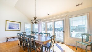 Un espace repas comprenant une grande table et des chaises, de grandes fenêtres équipées de stores et un lustre.
