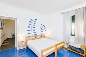 A bedroom with a bed, a bedside table, a small stool, a window with curtains, and a wall decoration. - Apartments Eos & Vespera (Dubrovnik)