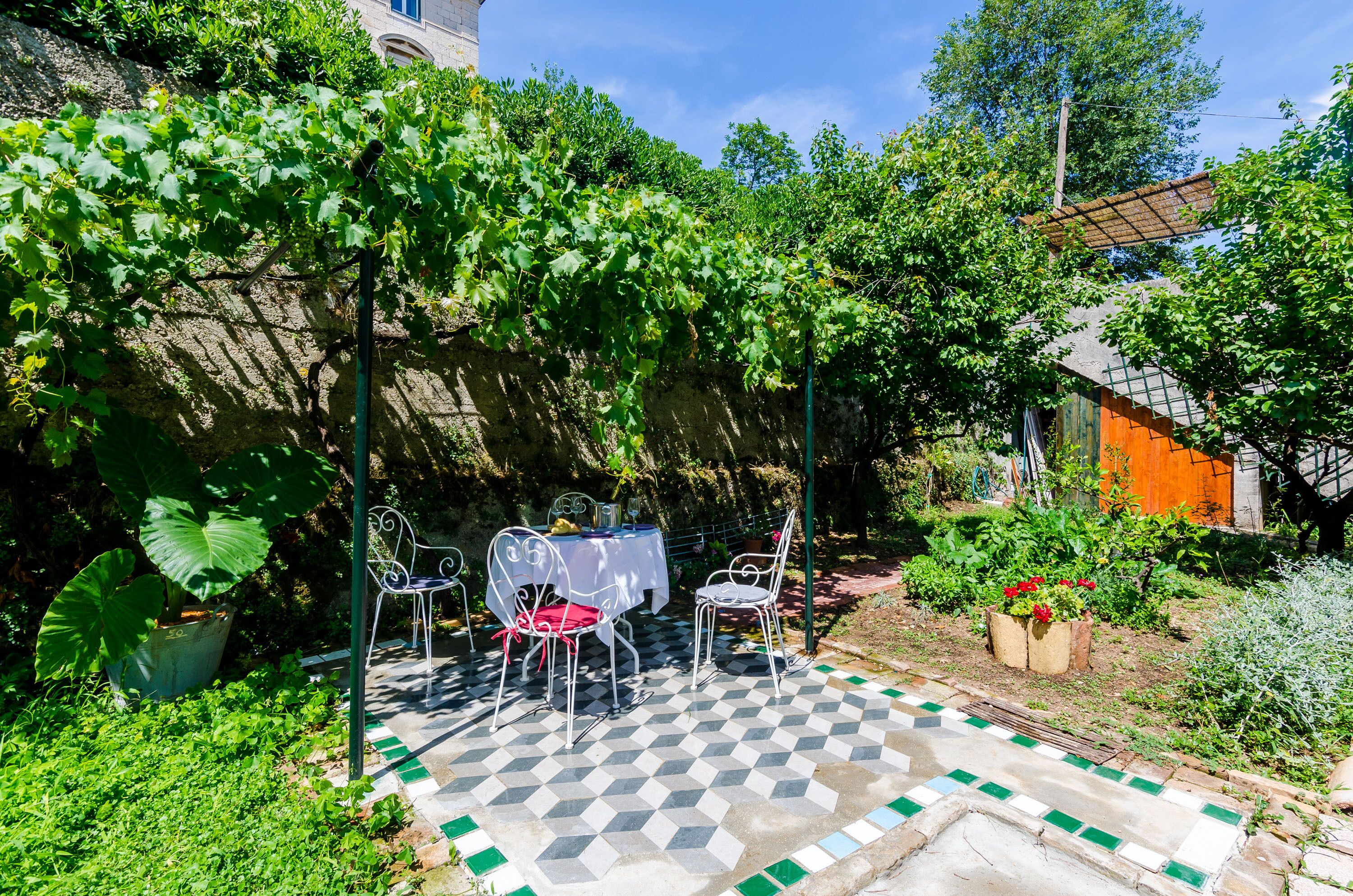 Une terrasse avec un sol à carreaux, deux chaises en métal blanc et une table recouverte d’une nappe blanche.