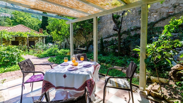 A covered outdoor dining area with a table set for two, surrounded by chairs, and a view of a lush garden and rocky hillside in the background. - Apartments Eos & Vespera (Dubrovnik)