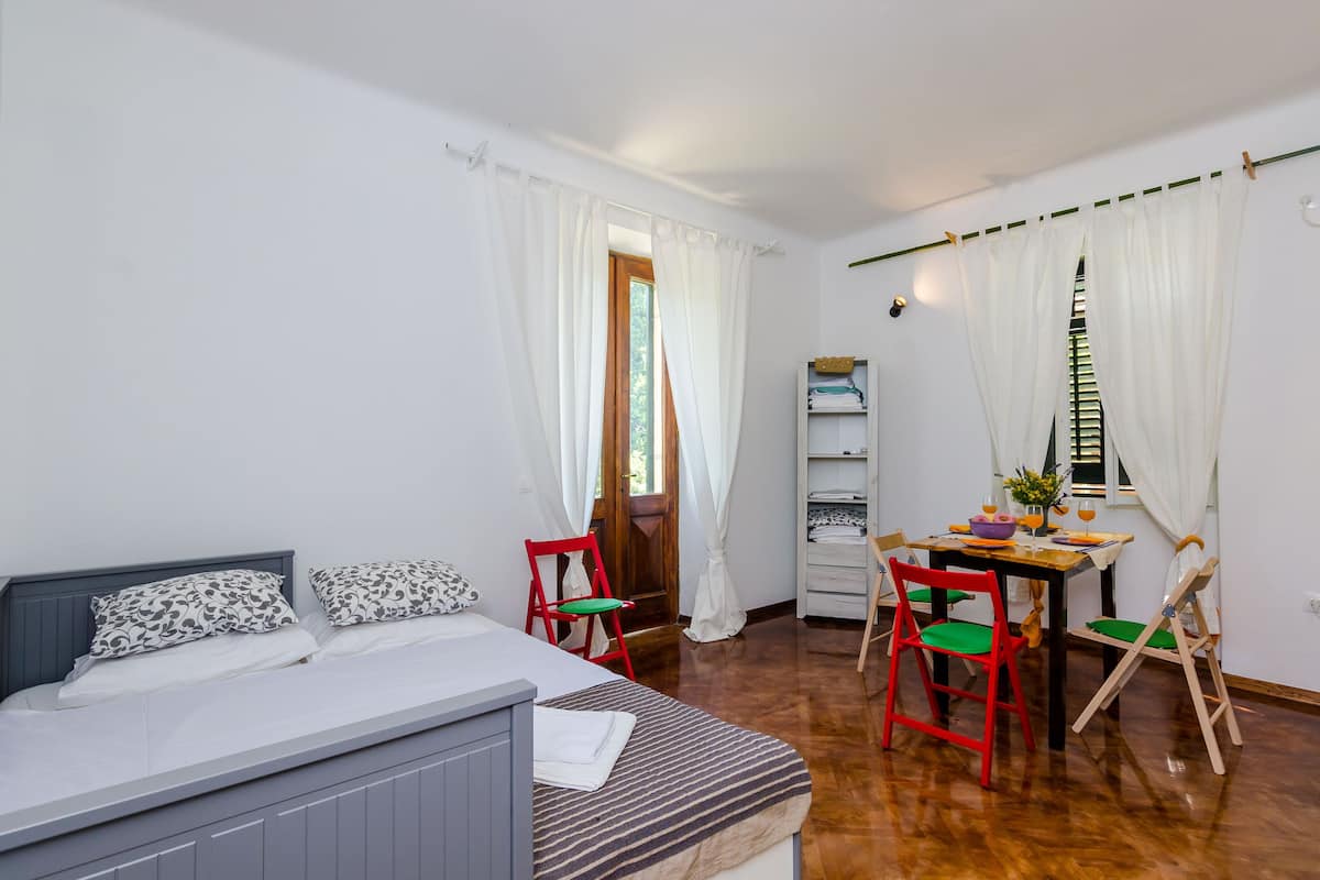 A bedroom with a bed, a small table with chairs, a wardrobe, and a window with curtains.