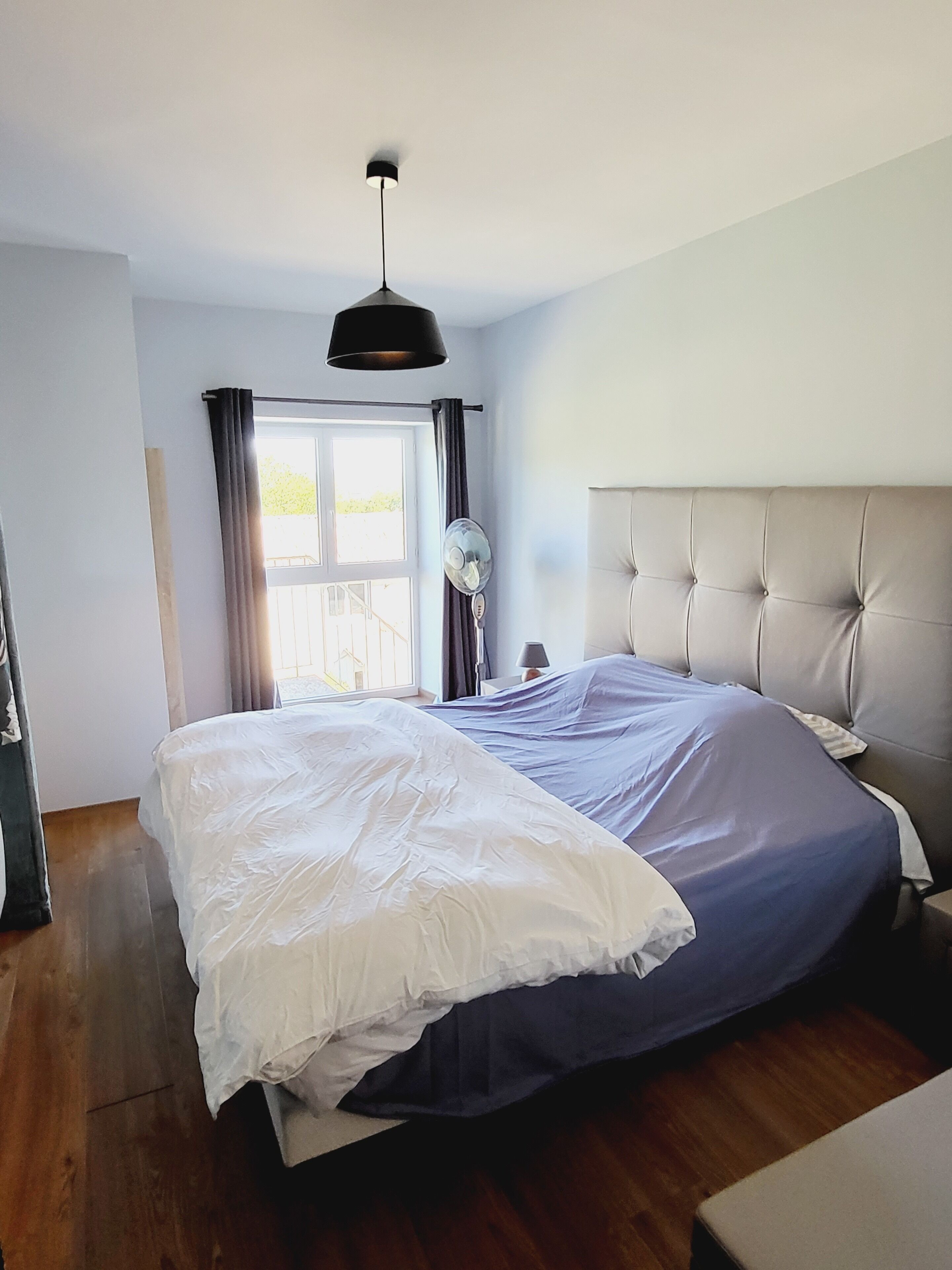 A bedroom with a large bed, a ceiling fan, a window with curtains, and a bedside table.