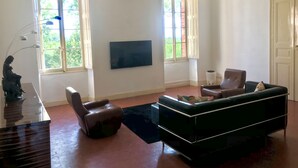 A living room with a black sofa, a brown armchair, a glass coffee table, and a TV mounted on the wall. There are two windows with white frames and a door on the right. - Gîte au Château Jonquier 2 (Séguret)