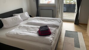 Cama doble con sábanas blancas, cabecera gris y vista a un paisaje urbano a través de una ventana con cortinas.