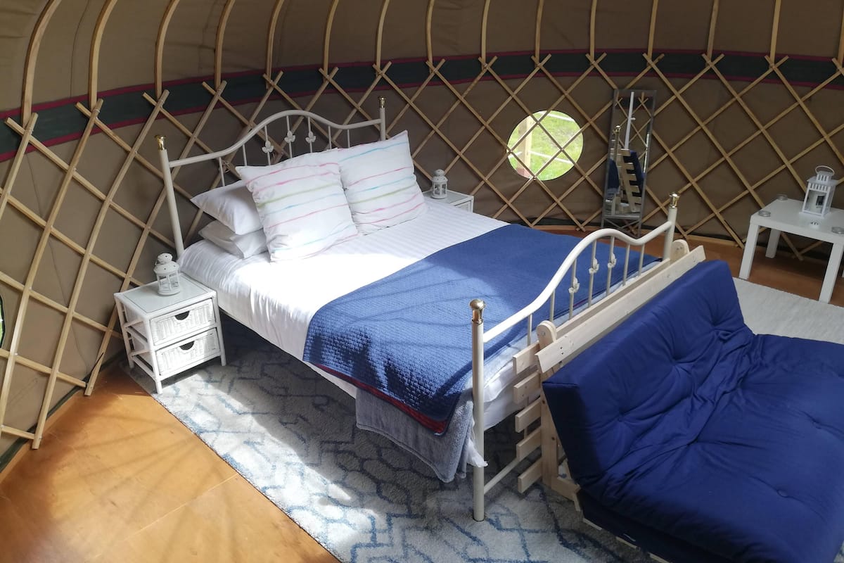 Un lit recouvert d’une couverture bleue et agrémenté d’oreillers blancs, dans une pièce de style yourte avec un plancher en bois.