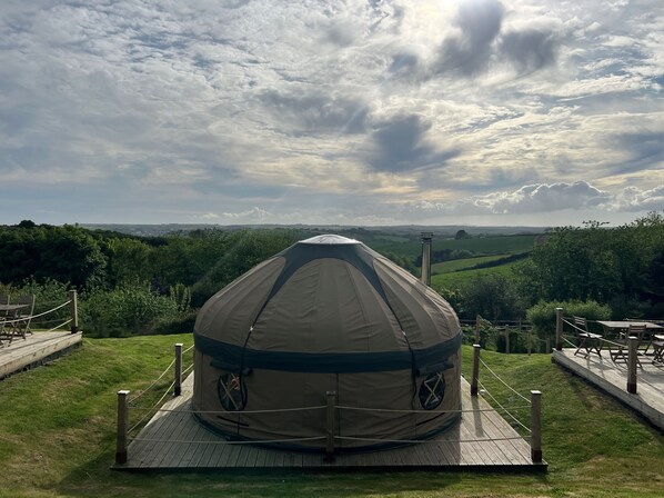 A large, dome-shaped tent with a circular entrance, surrounded by a wooden deck and metal railings, overlooking a green landscape with hills and a cloudy sky.