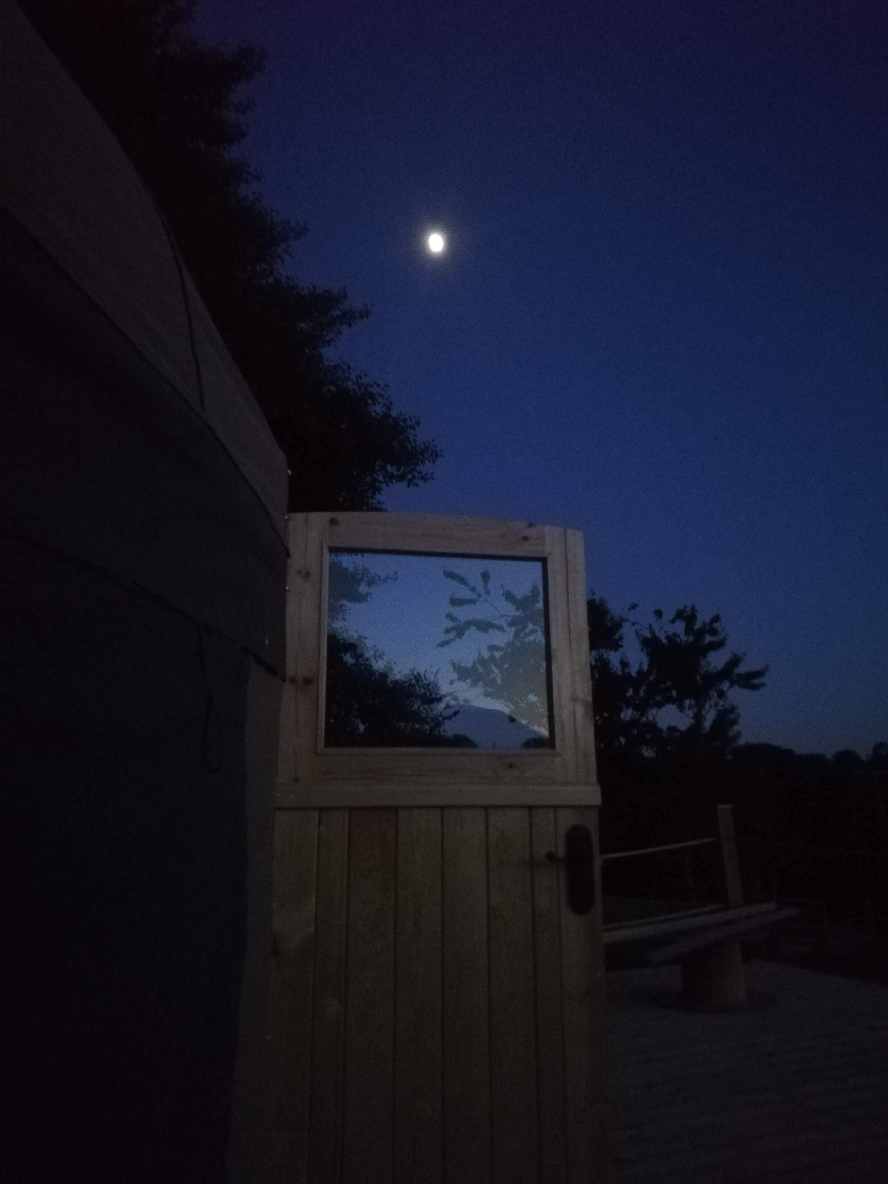 A wooden structure with a window reflecting trees and a moon in the sky.