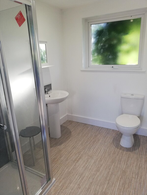 Baño con cabina de ducha de vidrio, lavabo, inodoro y ventana con vista a follaje.