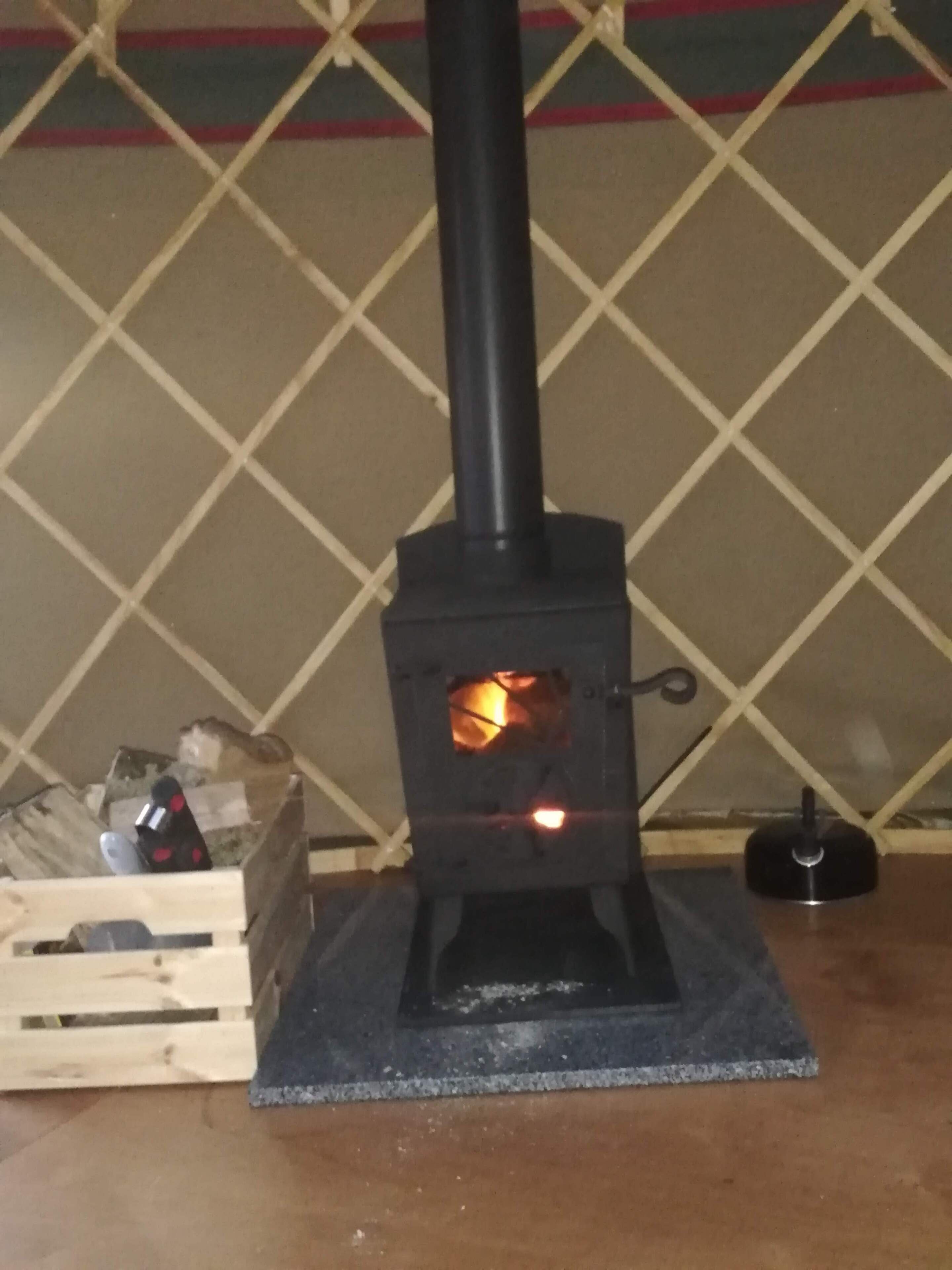 A wood-burning stove with a chimney, a wooden crate with items, and a metal kettle on a table.