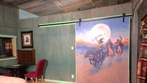 Un dormitorio con un amplio ventanal corredera que muestra un mural de vaqueros a caballo, una silla roja, una cama y una mesita con una taza.
