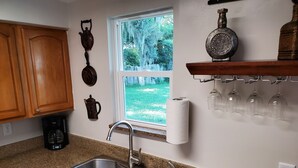 Une cuisine équipée d’un évier à deux bacs, de meubles en bois, d’une machine à café, d’une fenêtre donnant sur un espace verdoyant et d’une étagère où sont rangés des verres à vin.