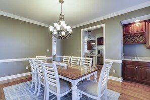 A dining room with a wooden table and white chairs, a chandelier, and a kitchen with wooden cabinets and stainless steel appliances.
