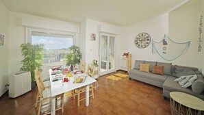 A spacious living room with a dining area, a gray sofa, and a view through a large window. - Petronella - Gardagate (Soiano)