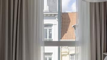 Habitació d'hotel amb una finestra gran, un llit amb roba de llit blanca, una manta blava a quadres i una bossa marró sobre el llit.
