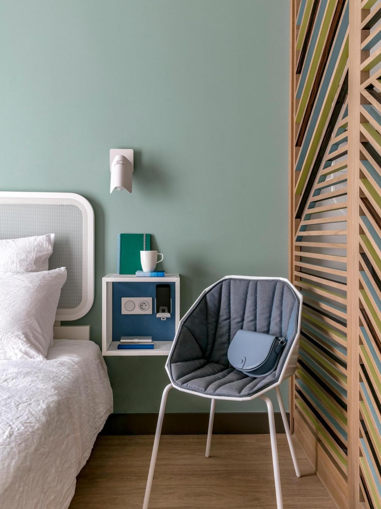 A bedroom with a bed, a chair, a nightstand, and a wall-mounted light.