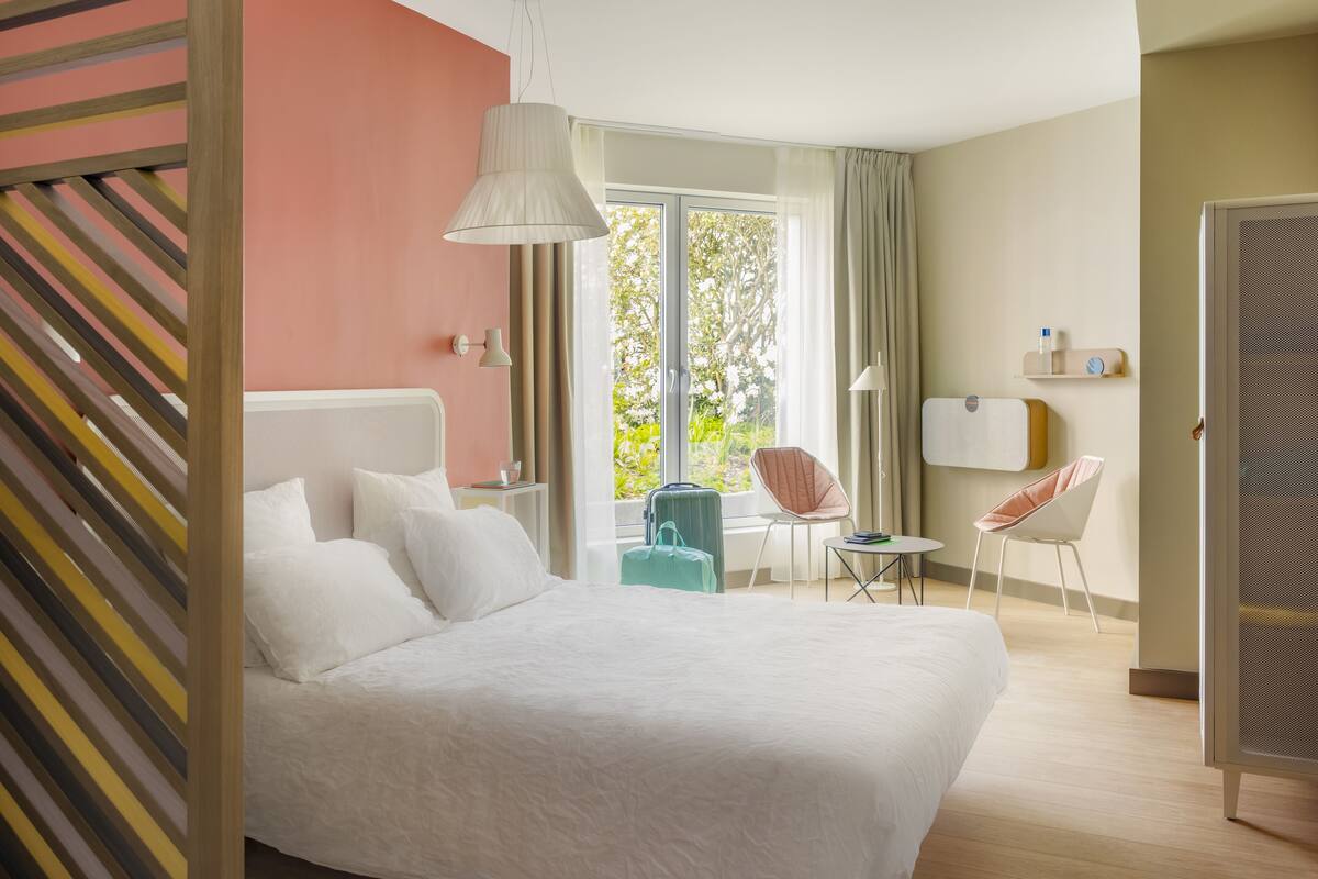 Habitación de hotel con una cama grande, un escritorio, dos sillas, un ventanal con vistas a zonas verdes y una puerta corredera.