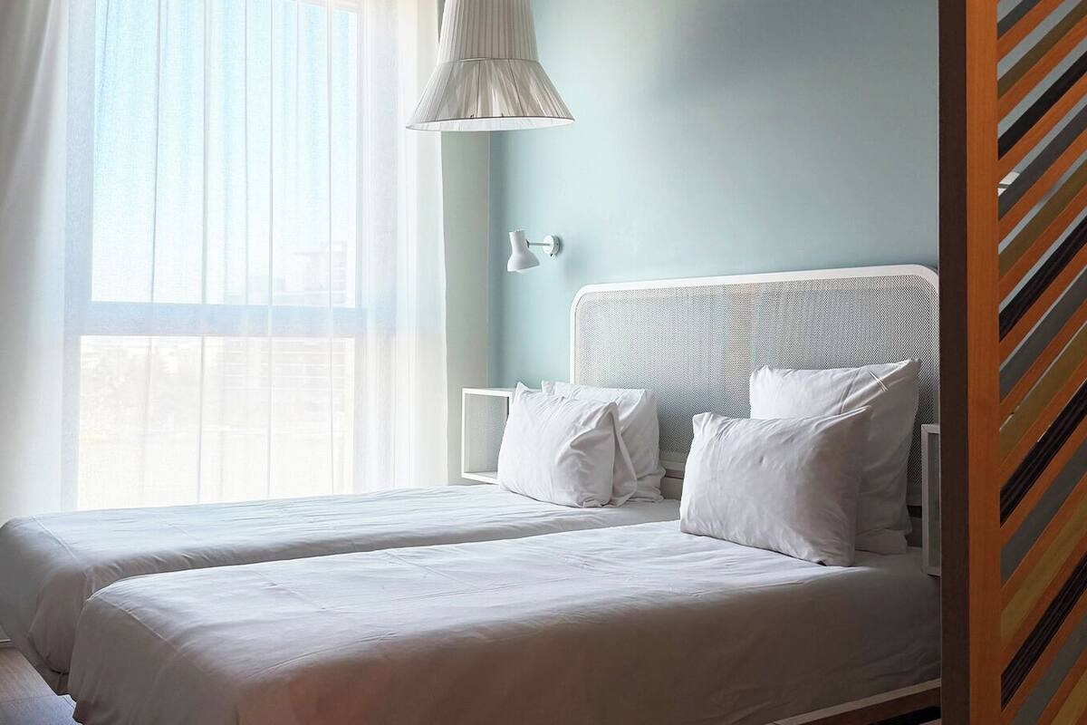 Habitación de hotel con dos camas, una ventana con cortinas, una lámpara blanca y una pared azul claro.