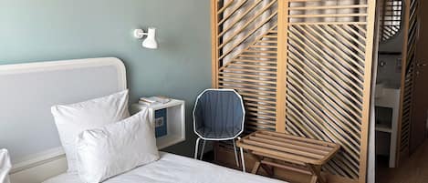 Habitación de hotel con cama, silla, mesita de noche, lámpara de pared y panel divisor de madera.