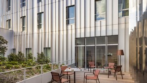 A modern outdoor seating area with a wooden deck, metal railings, and a glass building facade in the background. - OKKO Hotels Lille Centre (Lille)