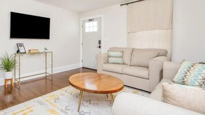Un salon avec un canapé beige, une table basse ronde en bois, un téléviseur fixé au mur et un luminaire suspendu au plafond.