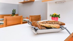 Ein Frühstücksbrett mit verschiedenen Brotsorten auf einem weißen Tisch in einem Essbereich mit Holzsesseln und einer Pflanze.