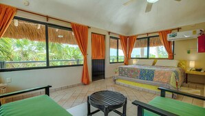 A room with a bed, a table, and chairs. There are windows with curtains, a ceiling fan, and a view of greenery outside.