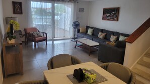 Ein Wohnzimmer mit einer Couch, Sesseln, einem Couchtisch und einem Esstisch. Ein Ventilator, eine Pflanze und Kunstwerke sind an den Wänden angebracht.