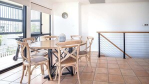 A dining area with a round table and six chairs, a clock on the wall, and a view through large windows.