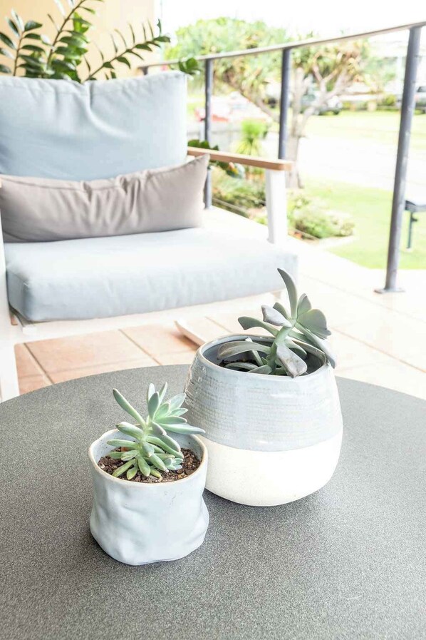 A patio with two potted plants on a table, a grey sofa with cushions, and a railing with greenery in the background.