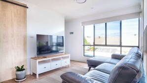 A modern living room with a flat-screen TV, a grey sofa, a white entertainment unit, and a large window offering a view of the outdoors.