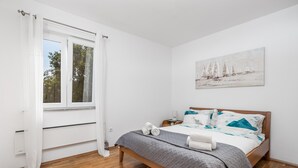 Une chambre avec un lit en bois, des murs blancs, une fenêtre avec des rideaux blancs, un tableau représentant des voiliers et un plancher en bois.