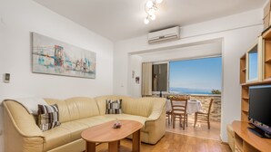 Un salon moderne avec un canapé beige, une table basse en bois et un téléviseur posé sur un meuble en bois. Un tableau orne le mur et, à travers une porte vitrée, on aperçoit la mer.