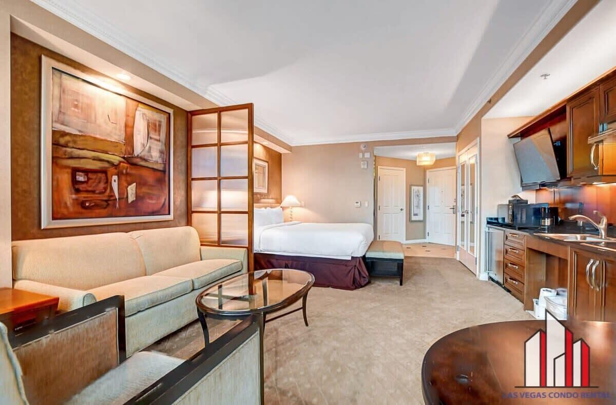 A hotel room with a sofa, a bed, a small table, a TV, and a kitchen area.