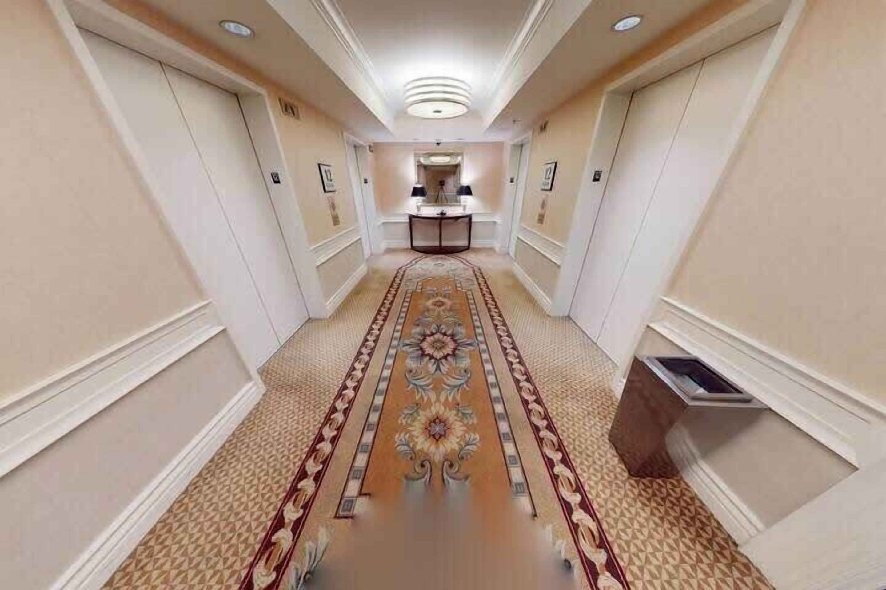 A hotel hallway with a central carpeted area, a small table with a lamp, and a trash bin.