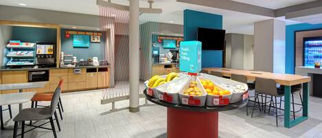 A modern cafeteria with a central fruit display, seating area, and a counter with food items.