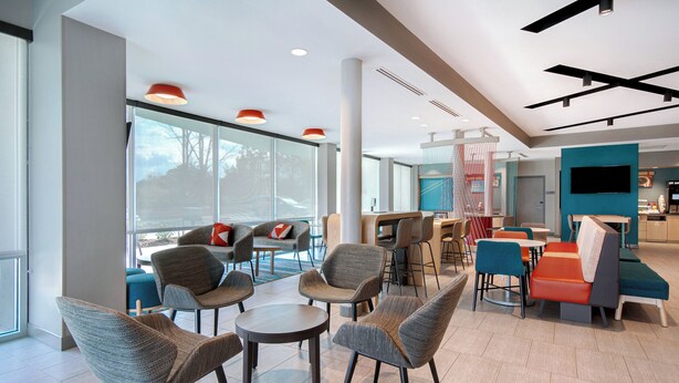 A modern hotel lobby with a seating area featuring round tables and chairs, a large TV screen, and a view of the outdoors. - avid Hotel Fort Mill - Carowinds Blvd by IHG (Fort Mill)