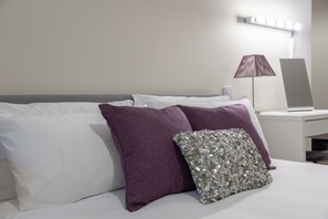 Una cama bien tendida con almohadas blancas y moradas, una almohada decorativa con lentejuelas y una mesita de noche con lámpara y espejo.