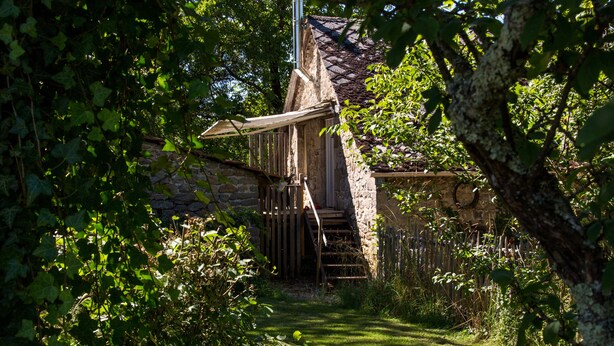 Ein Steinhaus mit einer Holztreppe, die zu einer Tür führt, umgeben von üppiger Vegetation und einem Holzzaun.