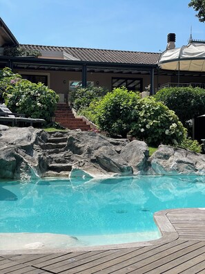 Uma área de piscina com deck de madeira, uma cascata de pedra e um edifício com telhado cerâmico ao fundo.