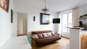 A cozy living room with a brown sofa, a white table, a TV, and a kitchen area with a coffee machine. - Beautiful flat in Montparnasse (Paris)