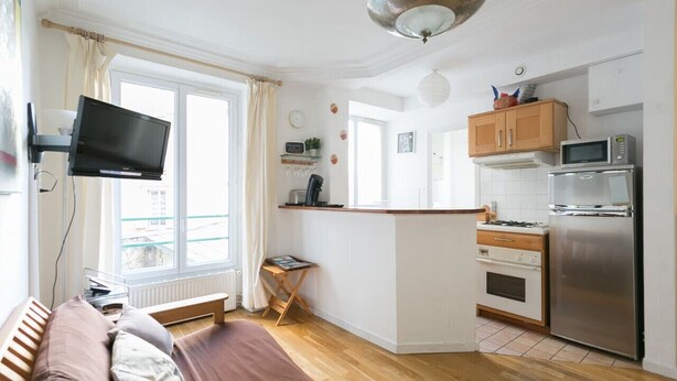 A compact living space with a kitchenette, a flat-screen TV mounted on the wall, a bed with a wooden headboard, and a window with curtains. - Beautiful flat in Montparnasse (Paris)