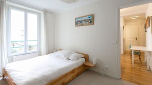 A bedroom with a wooden bed, a window with curtains, a painting on the wall, and a door leading to another room. - Beautiful flat in Montparnasse (Paris)