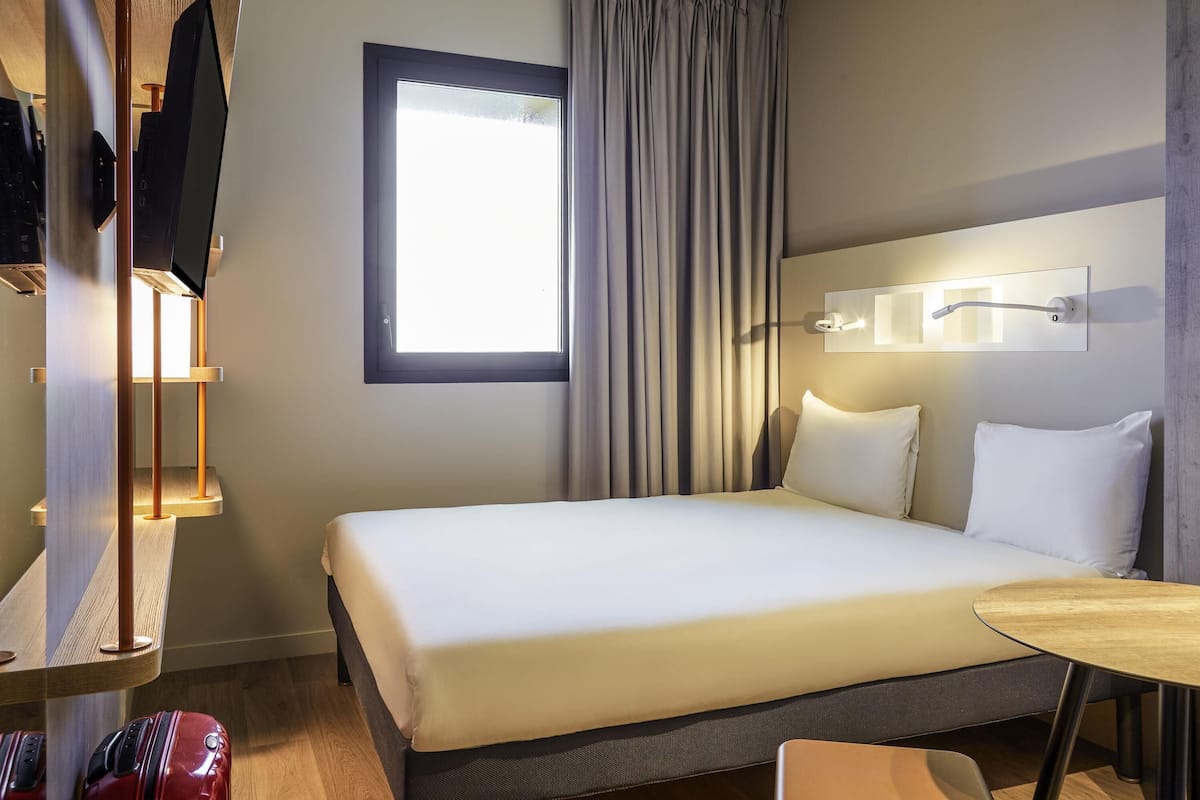 Ein Hotelzimmer mit Bett, Schreibtisch, Stuhl, Fernseher, einem Fenster mit Vorhängen und einer Wandleuchte.