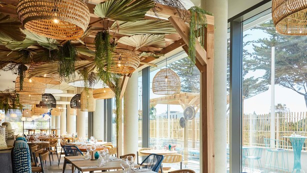 Un restaurant avec des tables et des chaises en bois, de grandes fenêtres et des lampes en osier suspendues au plafond.